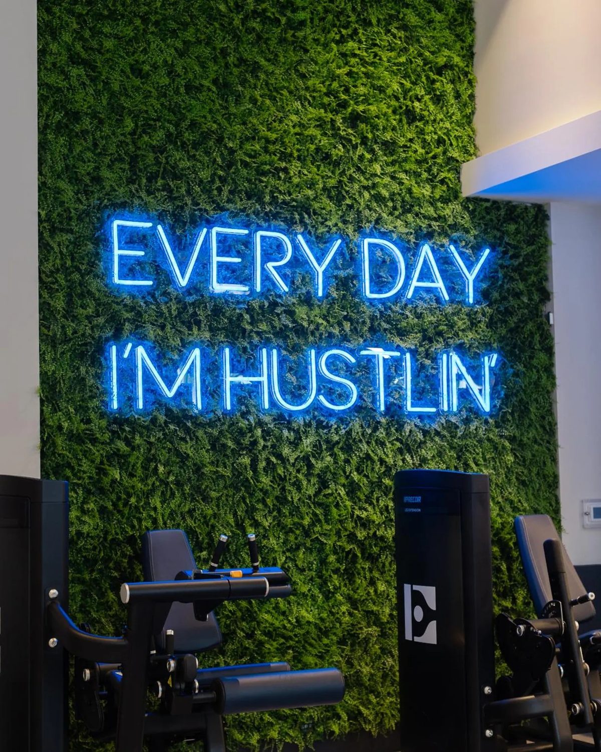 Неоновая надпись Every Day I'm Hustlin синим неоном на зелёной мховой стене в фитнес-зале