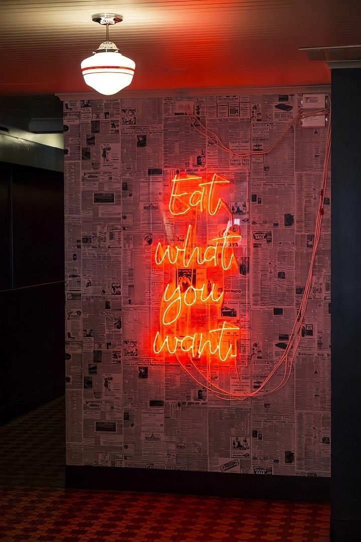 Неоновая надпись Eat What You Want оранжевым неоном на стене с газетными обоями